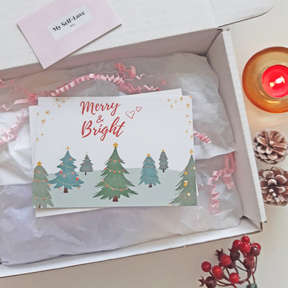 A Christmas greeting card with 'Merry & Bright' and a tree illustration, placed inside a white gift box with tissue paper and pink ribbon. A small card reading 'My Self-Love Box' is visible at the top, with a lit candle and pinecones around the box.