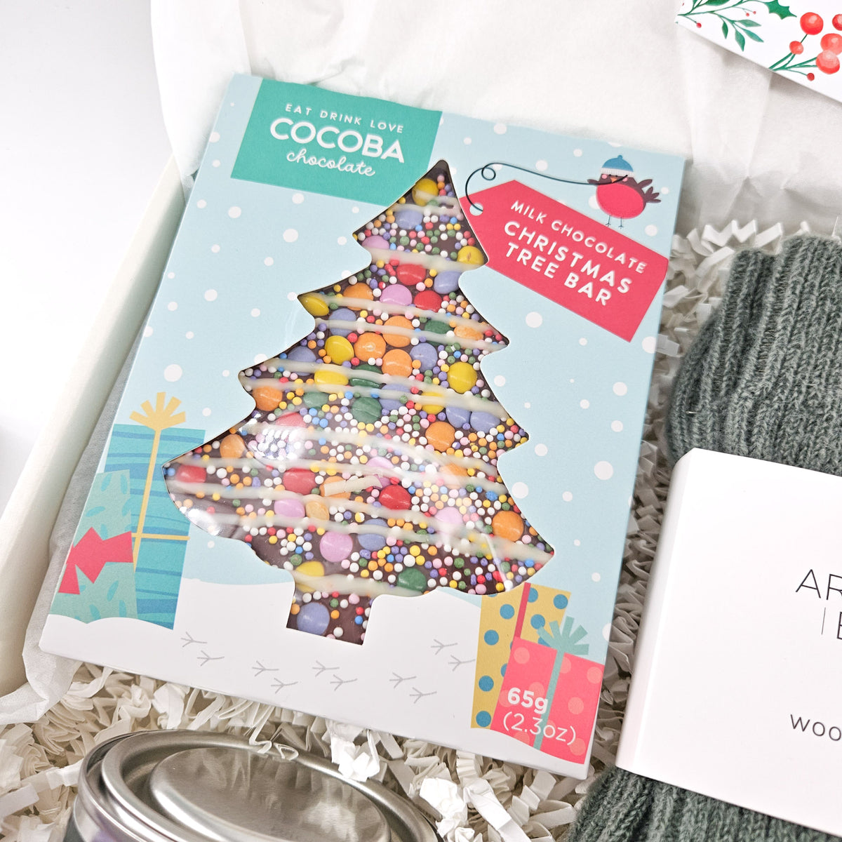 Close-up of Cocoba milk chocolate Christmas tree bar with colourful sweets, cosy grey wool socks and festive candle in a Christmas gift box.